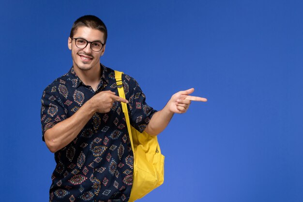 Vista frontale di uno studente maschio in camicia scura che indossa uno zaino giallo e sorridente sulla parete blu