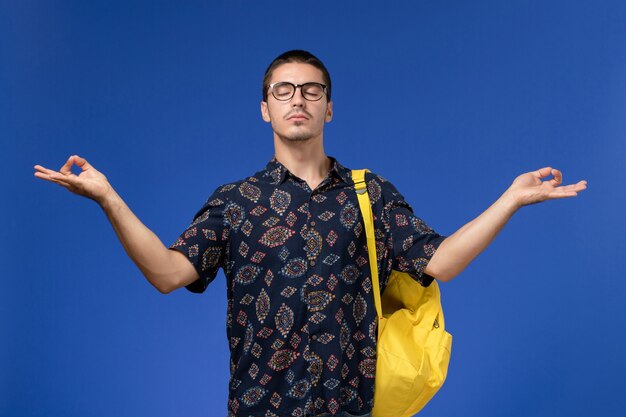 Vista frontale di uno studente maschio in camicia scura che indossa uno zaino giallo nella posa di meditazione sulla parete blu