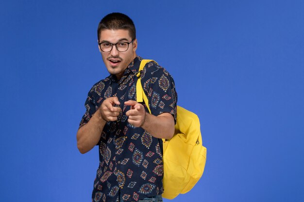 Vista frontale di uno studente maschio in camicia scura che indossa uno zaino giallo sulla parete blu chiaro