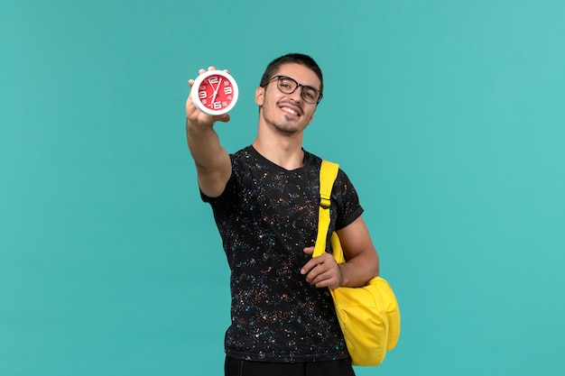 Vista frontale di uno studente maschio in t-shirt scura zaino giallo che tiene gli orologi sulla parete blu