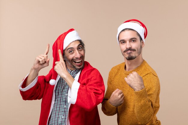 Vista frontale due amici con cappelli di Babbo Natale un dito del punto che mostra qualcosa in alto e l'altro che mostra i pugni su sfondo beige isolato