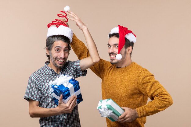 Vista frontale due ragazzi felici che tengono i regali di Natale e uno che toglie il cappello dell'altro su sfondo beige isolato