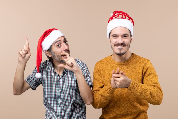 Vista frontale due ragazzi felici con cappelli di Babbo Natale uno che mostra qualcosa e l'altro tenendo le mani insieme su sfondo beige isolato