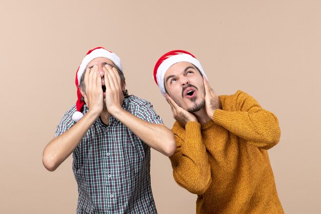 Vista frontale due ragazzi spaventati con cappelli di Babbo Natale uno che copre il viso e l'altro le orecchie su sfondo beige isolato