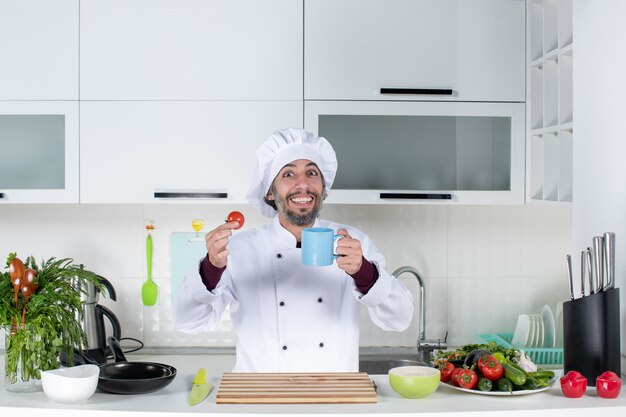 Vista frontale felice chef maschio con cappello da cuoco in piedi dietro il tavolo della cucina che sorregge tazza e pomodoro