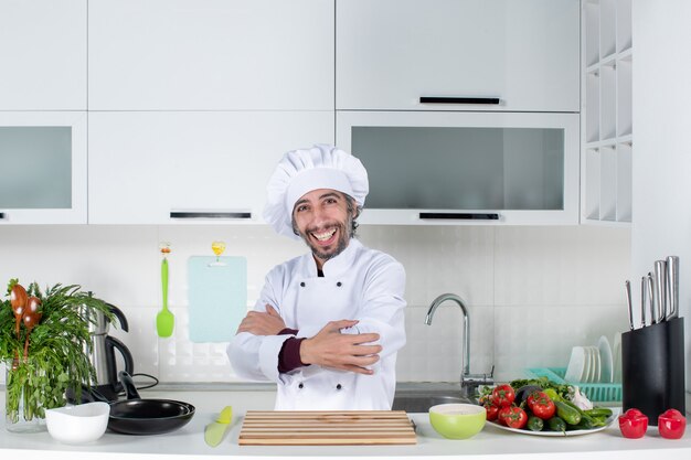 Vista frontale felice chef maschio in cappello da cuoco che incrocia le mani in piedi dietro il tavolo della cucina