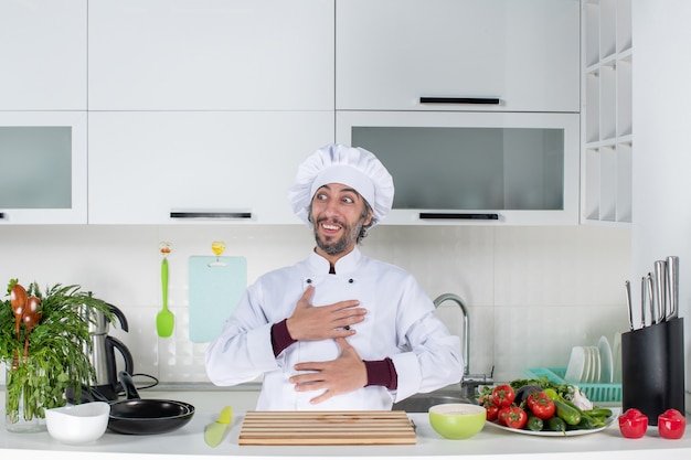 Vista frontale felice chef maschio in cappello da cuoco che si mette le mani sul petto in piedi dietro il tavolo della cucina