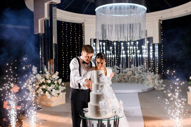 Vista frontale felice coppia sposata amorevole uomo e donna che tagliano una grande torta bianca sulla cerimonia di matrimonio di notte in piedi vicino all'altare di lusso ben decorato con fiori e stelle filanti Evento speciale