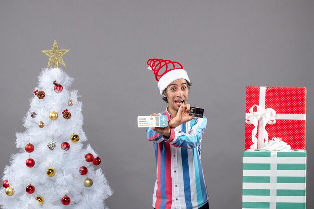 Vista frontale felice giovane uomo in possesso di carta e biglietto di viaggio con le mani incrociate intorno all'albero di Natale e regali