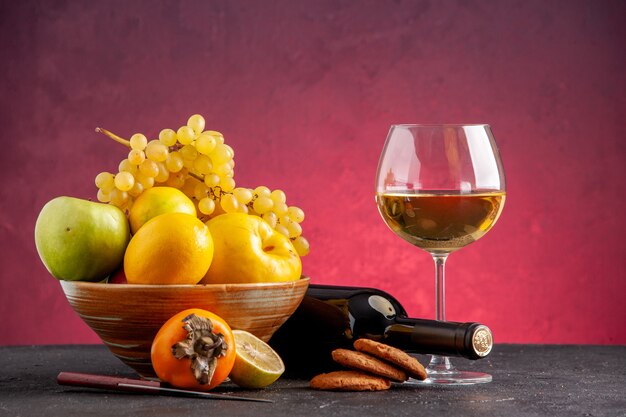 Vista frontale frutta fresca in una ciotola di legno mela mela cotogna uva limone cachi bottiglia di vino rovesciata bicchiere di vino biscotti sul tavolo rosso