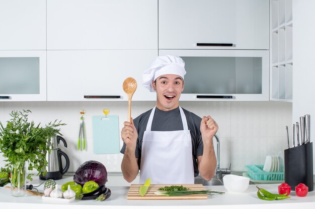 Vista frontale gioioso chef maschio in uniforme che tiene un cucchiaio di legno dietro il tavolo della cucina