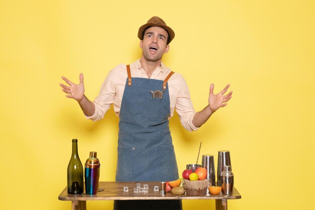 Vista frontale giovane barista maschio davanti al tavolo con bevande shaker su sfondo giallo