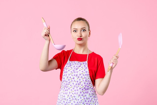 Vista frontale giovane bella casalinga in mantello che tiene cucchiai sul muro rosa
