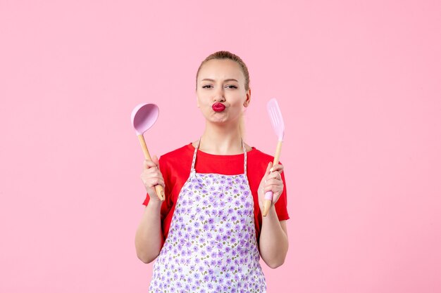 Vista frontale giovane bella casalinga in mantello con cucchiai sul muro rosa