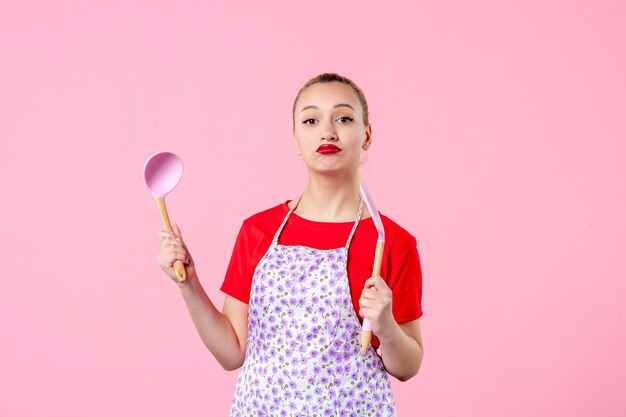 Vista frontale giovane bella casalinga in mantello con cucchiai sul muro rosa