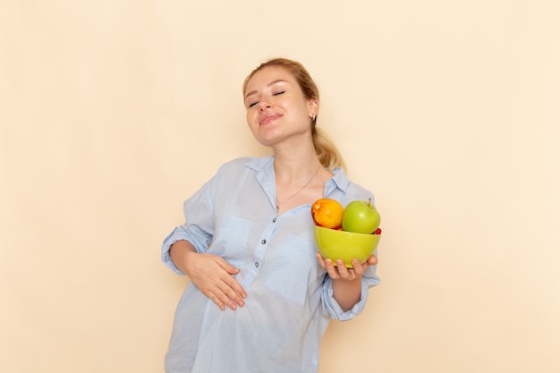 Vista frontale giovane bella femmina in camicia tenendo il piatto con frutta e sorridente in posa sulla parete crema modello di frutta donna posa signora