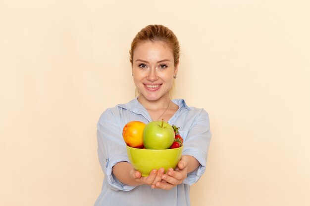 Vista frontale giovane bella femmina in camicia tenendo il piatto con frutta e sorridente sulla parete crema modello di frutta donna posa signora