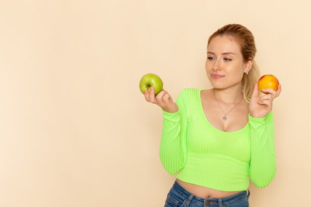 Vista frontale giovane bella femmina in camicia verde che tiene un paio di mele sulla parete crema frutta modello donna mellow