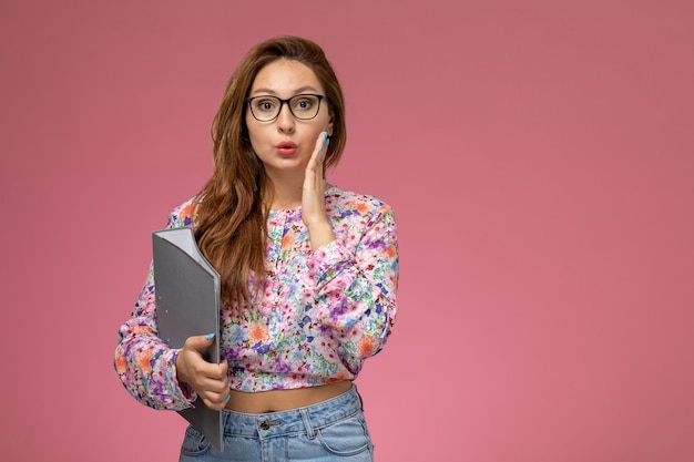 Vista frontale giovane bella femmina in fiore progettato camicia e blue jeans che tengono documento grigio su sfondo rosa