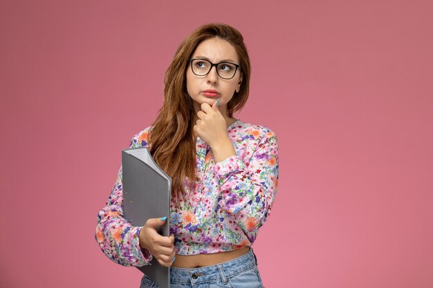 Vista frontale giovane bella femmina in fiore progettato camicia e blue jeans che tengono documento grigio su sfondo rosa