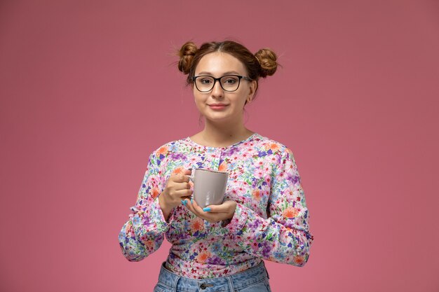 Vista frontale giovane bella femmina in fiore progettato camicia e blue jeans che tengono tazza di tè con il sorriso su sfondo rosa