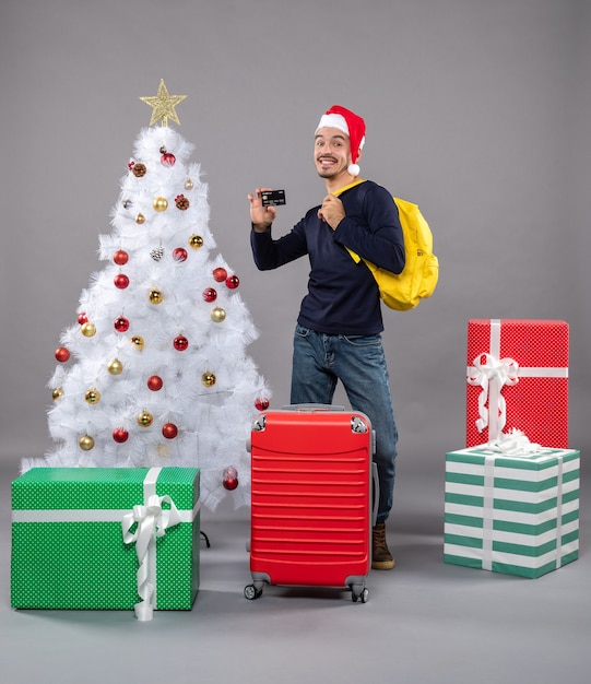 Vista frontale giovane con cappello da Babbo Natale e valigia rossa che mostra una carta su grigio isolato