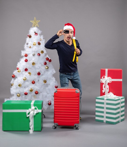 Vista frontale giovane con carta e valigia rossa in piedi vicino all'albero di Natale e regali su grigio isolato