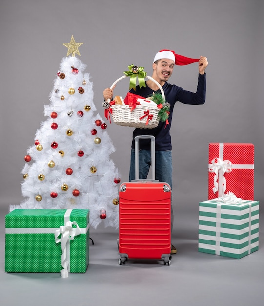 Vista frontale giovane con cesto regalo e cappello da Babbo Natale vicino all'albero di Natale su grigio isolato