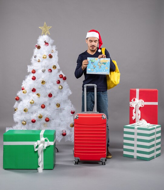 Vista frontale giovane con zaino giallo che tiene e guarda la mappa vicino all'albero di Natale e presenta su grigio isolato