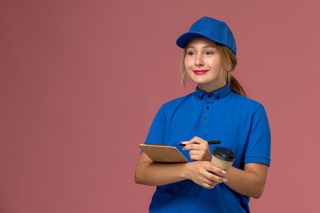 Vista frontale giovane corriere femminile in uniforme blu in posa tenendo la tazza di caffè e blocco note con un leggero sorriso, donna di consegna uniforme di servizio