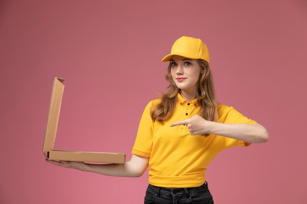 Vista frontale giovane corriere femminile in uniforme gialla che tiene scatola di cibo marrone aprendolo sulla lavoratrice servizio di consegna uniforme scrivania rosa scuro