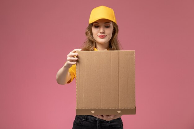 Vista frontale giovane corriere femminile in uniforme gialla che tiene scatola di cibo marrone aprendolo sulla lavoratrice servizio di consegna uniforme scrivania rosa scuro