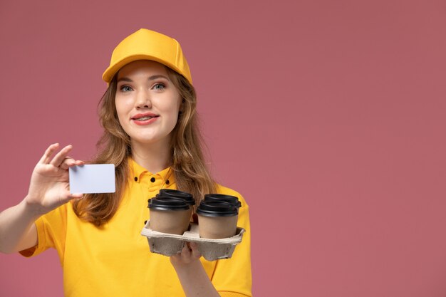 Vista frontale giovane corriere femminile in uniforme gialla che tiene tazze di caffè con carta bianca sulla lavoratrice servizio di consegna uniforme scrivania rosa scuro