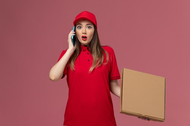 Vista frontale giovane corriere femminile in uniforme rossa che tiene la scatola di cibo per la consegna mentre parla al telefono su sfondo rosa chiaro società uniforme di servizio di consegna