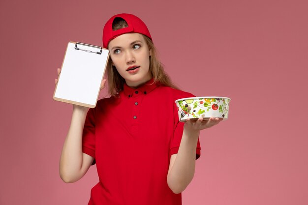 Vista frontale giovane corriere femminile in uniforme rossa e mantello che tiene la ciotola di consegna e il blocco note che pensa sulla parete rosa