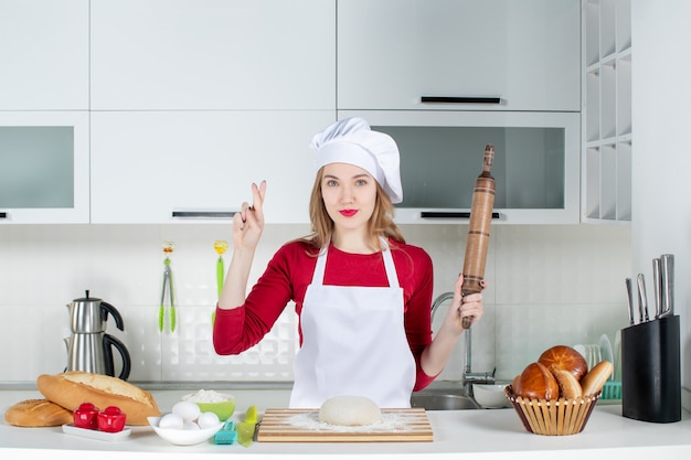 Vista frontale giovane cuoca che tiene il mattarello facendo segno di buona fortuna in cucina