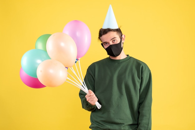 Vista frontale giovane deluso con cappello da festa e palloncini colorati in piedi sul giallo