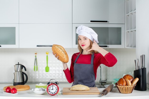 Vista frontale giovane donna con cappello da cuoco e grembiule che fa un cartello okey che tiene il pane in cucina