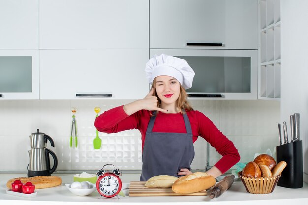 Vista frontale giovane donna con cappello da cuoco e grembiule che mi fa un gesto telefonico in cucina