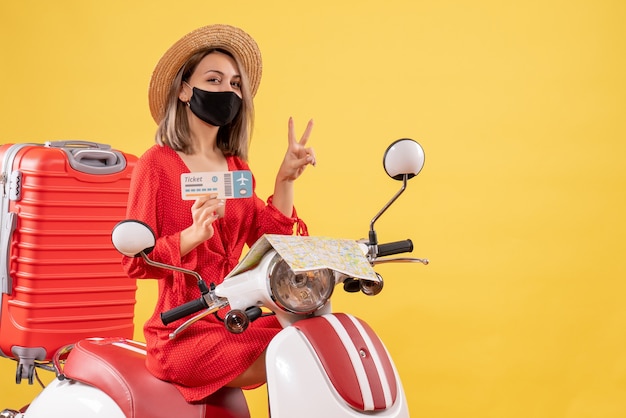 Vista frontale giovane donna con maschera nera su ciclomotore con valigia rossa che tiene biglietto che gesturing segno di vittoria