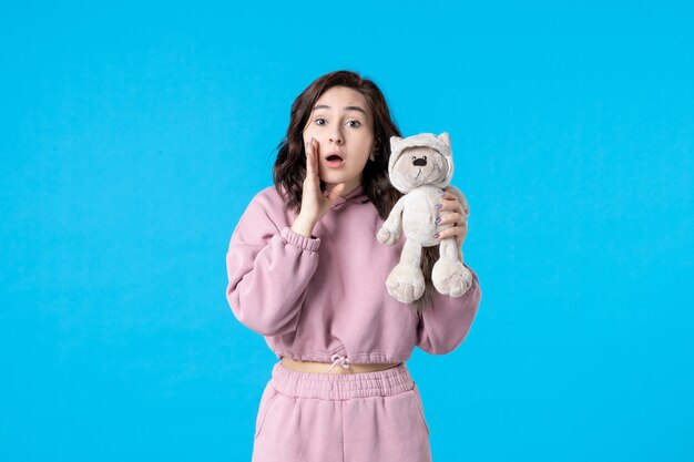 Vista frontale giovane donna in pigiama rosa con piccolo orso giocattolo sul colore blu