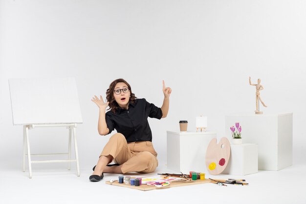 Vista frontale giovane donna seduta all'interno della stanza con cavalletto e vernici su scrivania bianca arte a colori disegnare pittore artista disegno