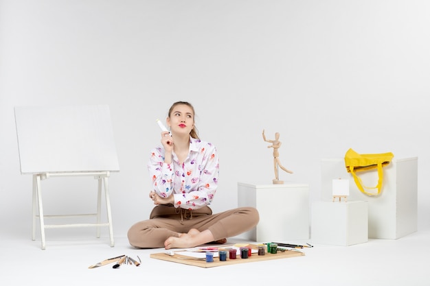 Vista frontale giovane donna seduta con vernici e cavalletto per disegnare su sfondo bianco