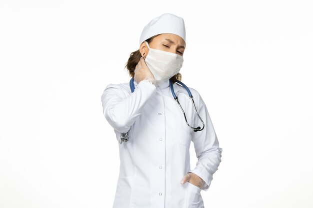 Vista frontale giovane dottoressa con maschera sterile e guanti a causa del coronavirus che ha mal di collo sulla superficie bianca