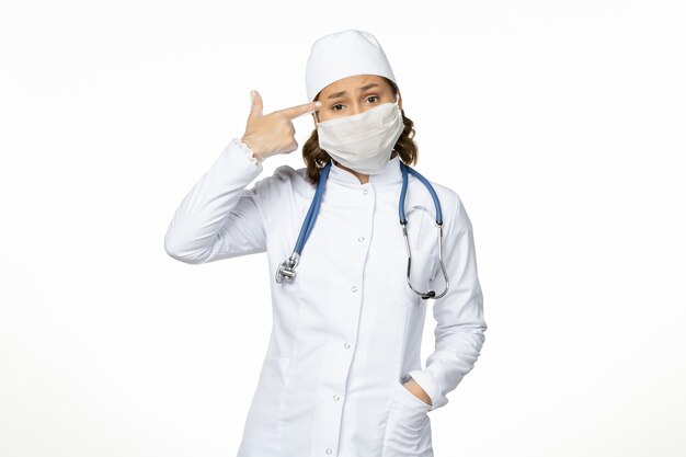 Vista frontale giovane dottoressa in tuta medica bianca e con maschera a causa del coronavirus sul pavimento bianco