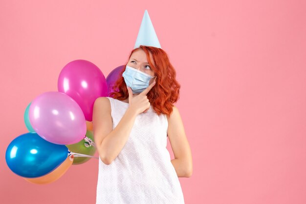 Vista frontale giovane femmina che nasconde palloncini colorati in maschera sterile pensando su sfondo rosa covid festa- colore di capodanno natale