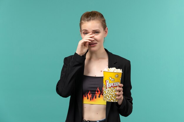 Vista frontale giovane femmina che tiene pacchetto di popcorn e guardare film sulla scrivania blu