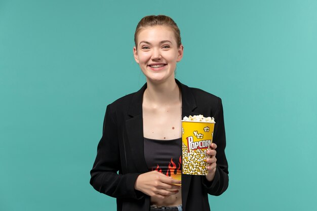 Vista frontale giovane femmina che tiene pacchetto di popcorn guardando film e ridendo sulla superficie blu