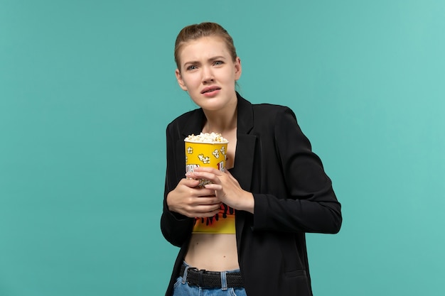 Vista frontale giovane femmina che tiene pacchetto di popcorn guardando film sulla superficie blu chiaro