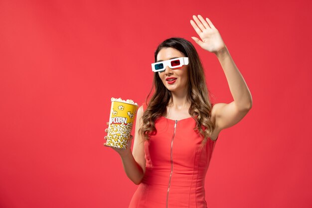 Vista frontale giovane femmina che tiene pacchetto di popcorn in occhiali da sole d sulla superficie rossa